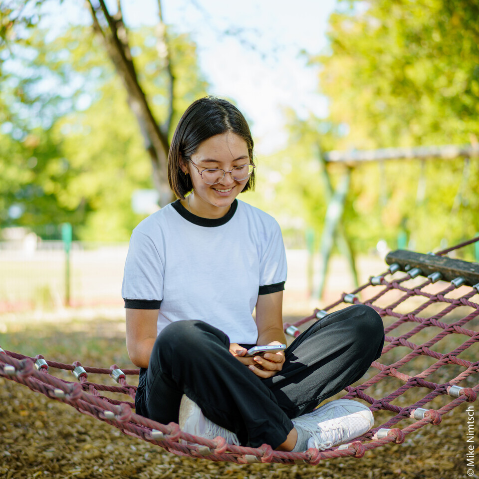 Eine junge Frau sitzt in der Hängematte und blickt auf ihr Telefon. | © Mike Nimtsch